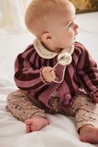 A baby sitting on white bedding, wearing a knitted burgundy and mauve striped cardigan with a cream floral collar and patterned pants, holding a silver bell.