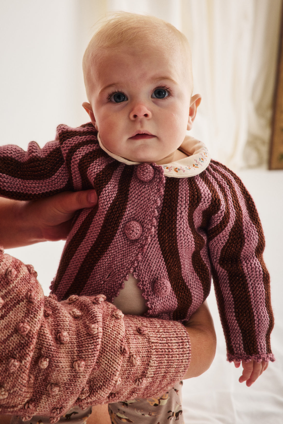 A close-up of a baby wearing a knitted cardigan with vertical stripes in dusty rose and dark brown, held by an adult wearing a matching textured knit sweater.