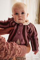 A close-up of a baby wearing a knitted cardigan with vertical stripes in dusty rose and dark brown, held by an adult wearing a matching textured knit sweater.