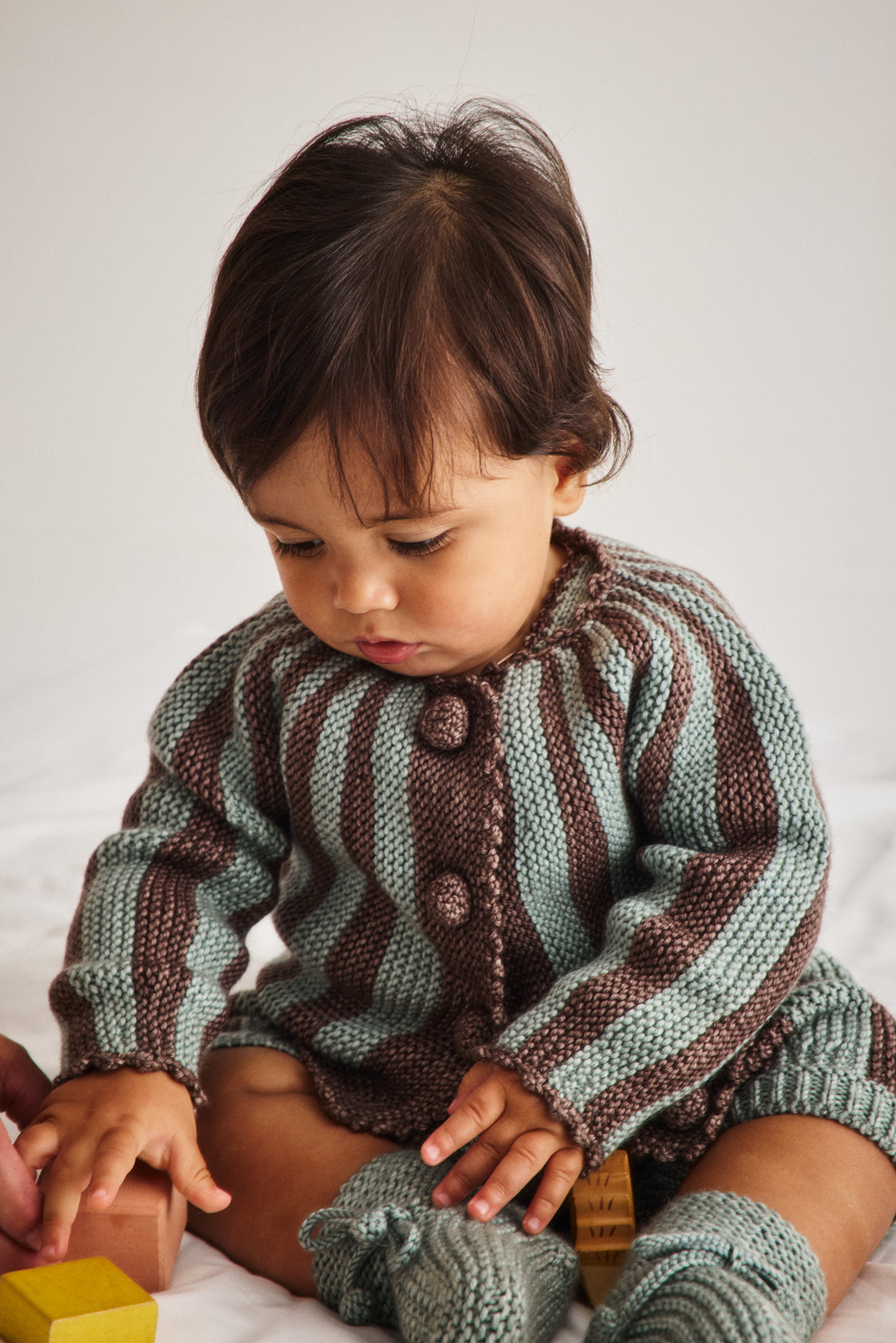 A baby wearing a knitted striped cardigan and booties in sage green and brown, sitting on a white surface and looking down at a wooden block.