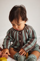 A baby wearing a knitted striped cardigan and booties in sage green and brown, sitting on a white surface and looking down at a wooden block.
