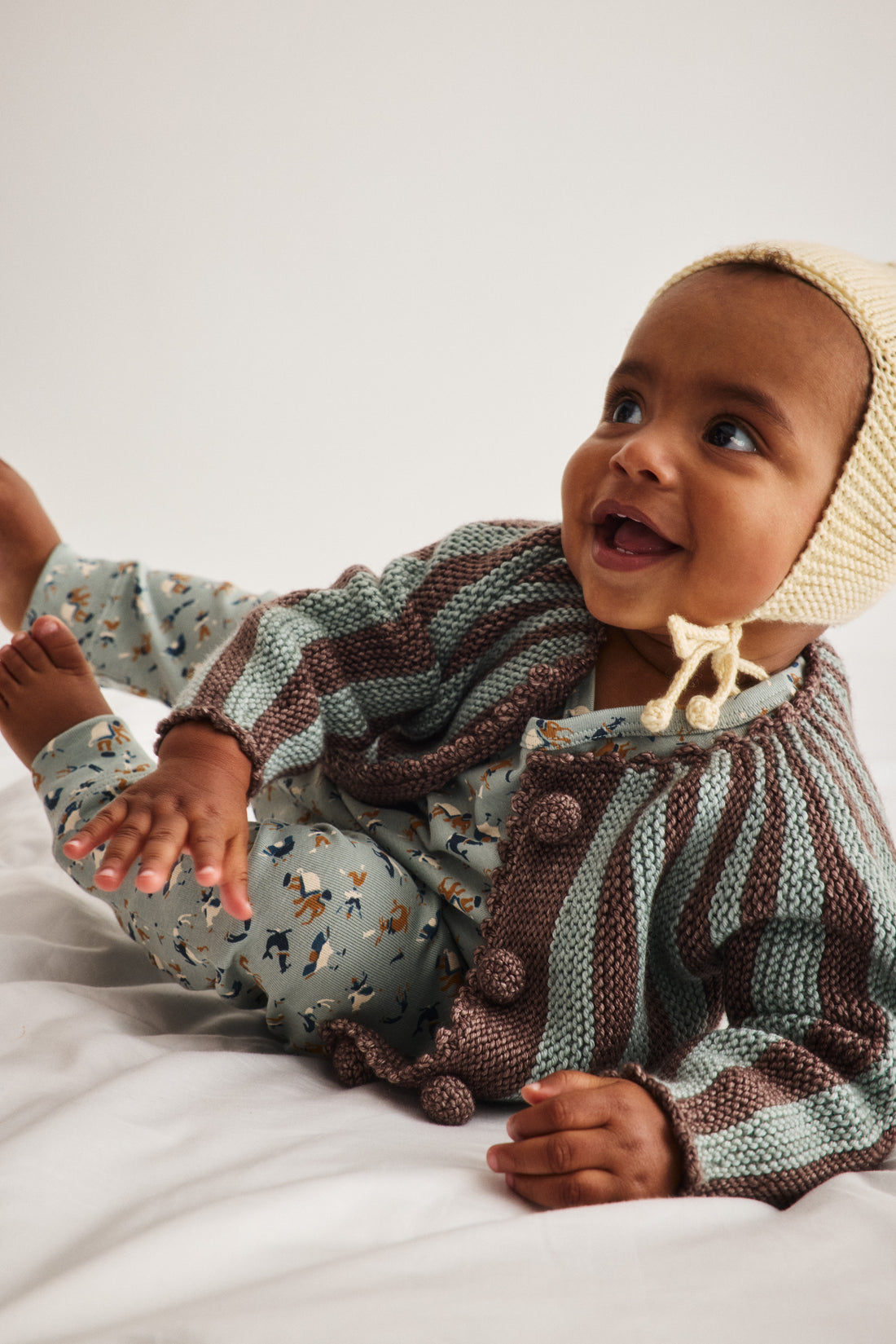 A baby wearing a knitted two-piece outfit with a brown and sage green striped cardigan and matching patterned pants, along with a cream bonnet, lying on a white bed.