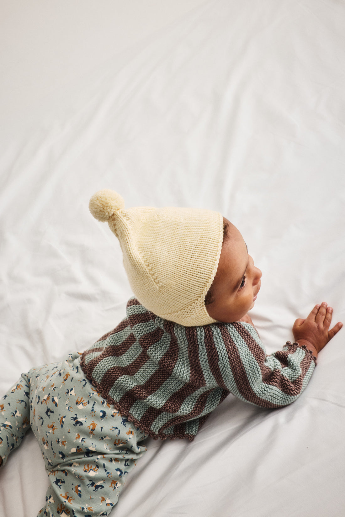 A baby lying on a white sheet, wearing a cream knit pom-pom beanie, a green and brown striped knit sweater, and light blue patterned pants.