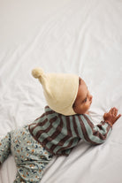 A baby lying on a white sheet, wearing a cream knit pom-pom beanie, a green and brown striped knit sweater, and light blue patterned pants.
