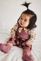 A baby sitting on white sheets wearing a matching knit outfit with a dusty rose pinafore-style romper, patterned cream long-sleeve shirt, and matching booties.