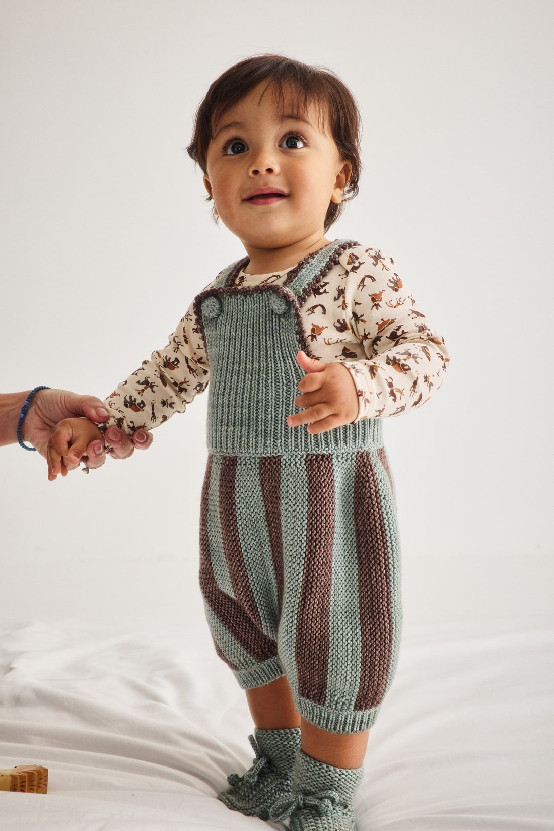 A baby wearing a knitted overall romper with vertical stripes in sage green and brown, layered over a long-sleeve cream shirt with a brown animal print, standing on a white blanket and holding an adult's hand.
