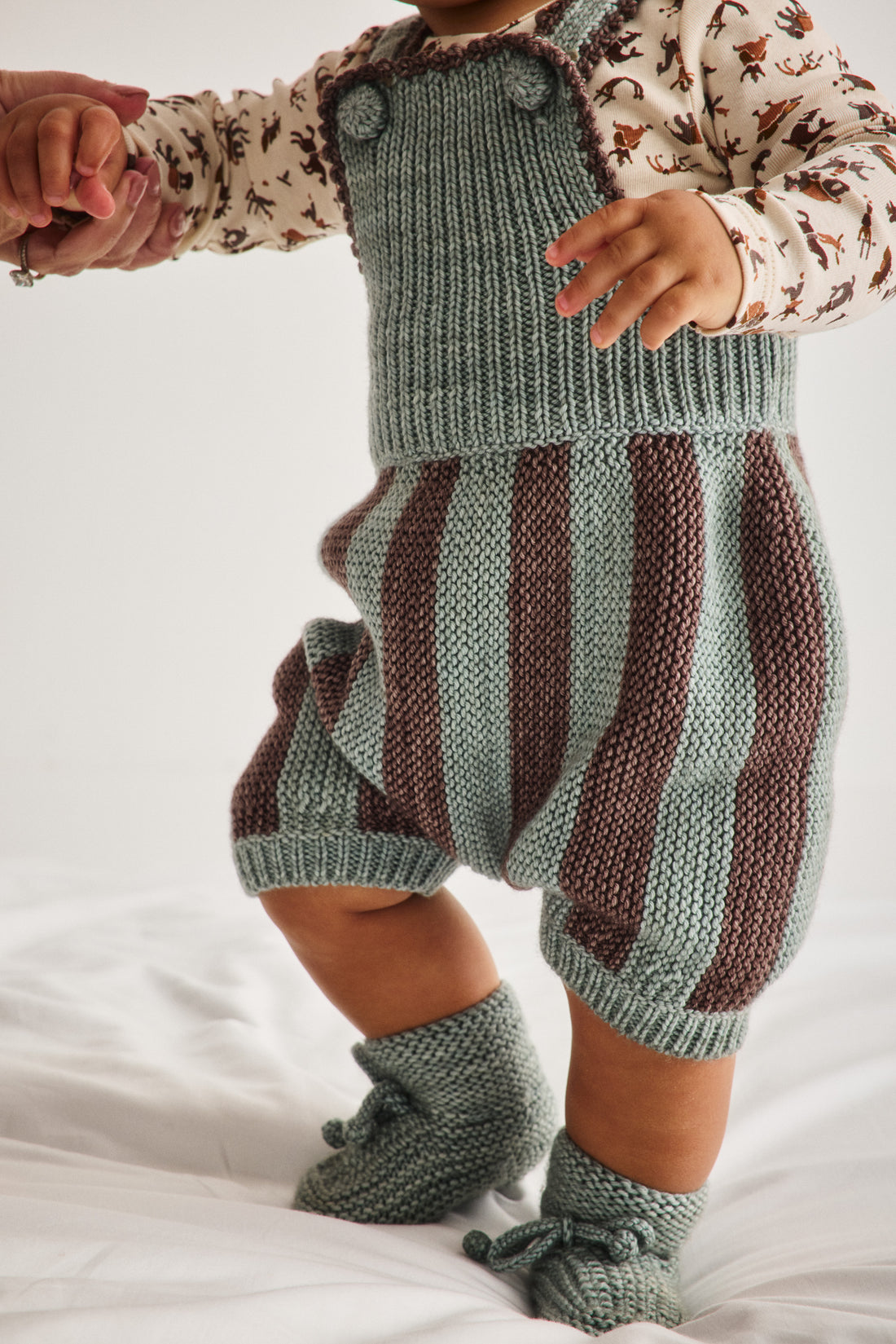 A baby wearing a knitted romper with dusty blue-green and dark brown vertical stripes, paired with matching knit booties and a patterned long-sleeve shirt, standing on a white fabric surface.