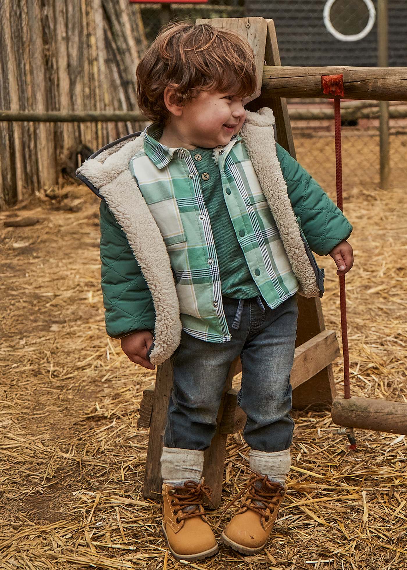 Child wearing a green plaid shirt, brown boots, and a puffer vest standing on straw.