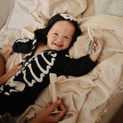 Baby wearing a skeleton outfit lying on a bed with a pacifier.