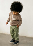 A toddler with a large black afro stands against a white wall wearing a taupe knit sweater with white dinosaur graphics, sage green pants, and navy sneakers.
