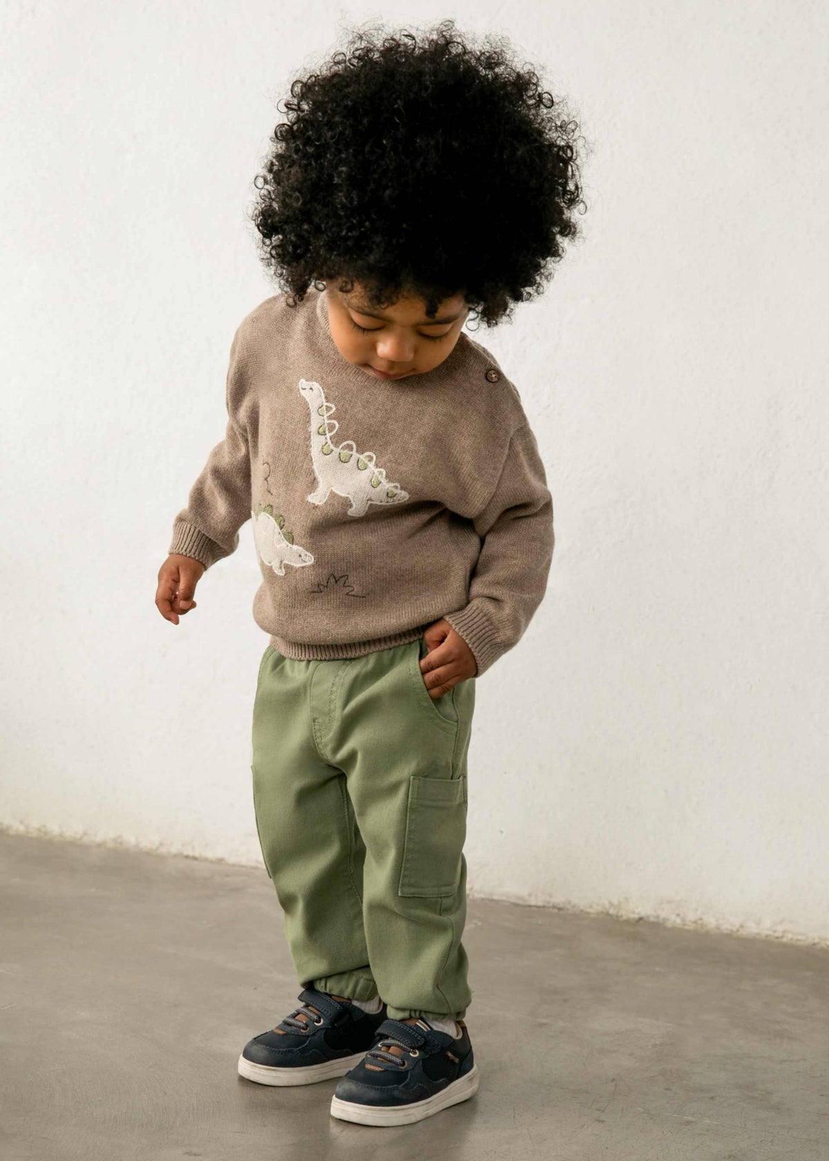 A toddler with a large black afro stands against a white wall wearing a taupe knit sweater with white dinosaur graphics, sage green pants, and navy sneakers.