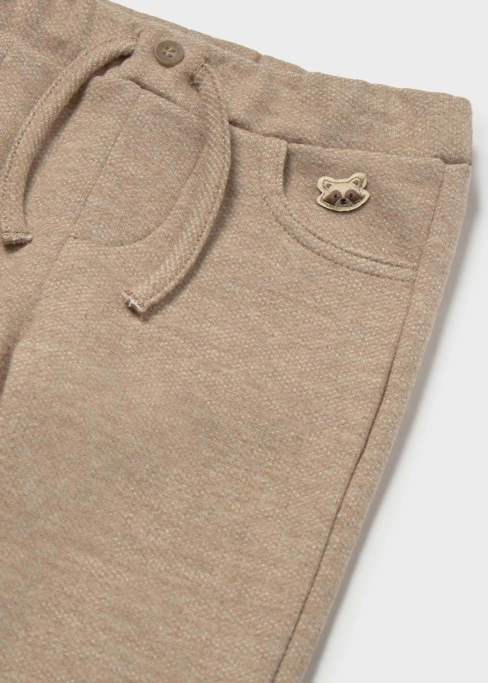 Close-up of beige heathered knit pants showing a drawstring waist, front pocket, and a small metallic raccoon logo on the pocket.