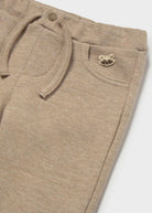 Close-up of beige heathered knit pants showing a drawstring waist, front pocket, and a small metallic raccoon logo on the pocket.