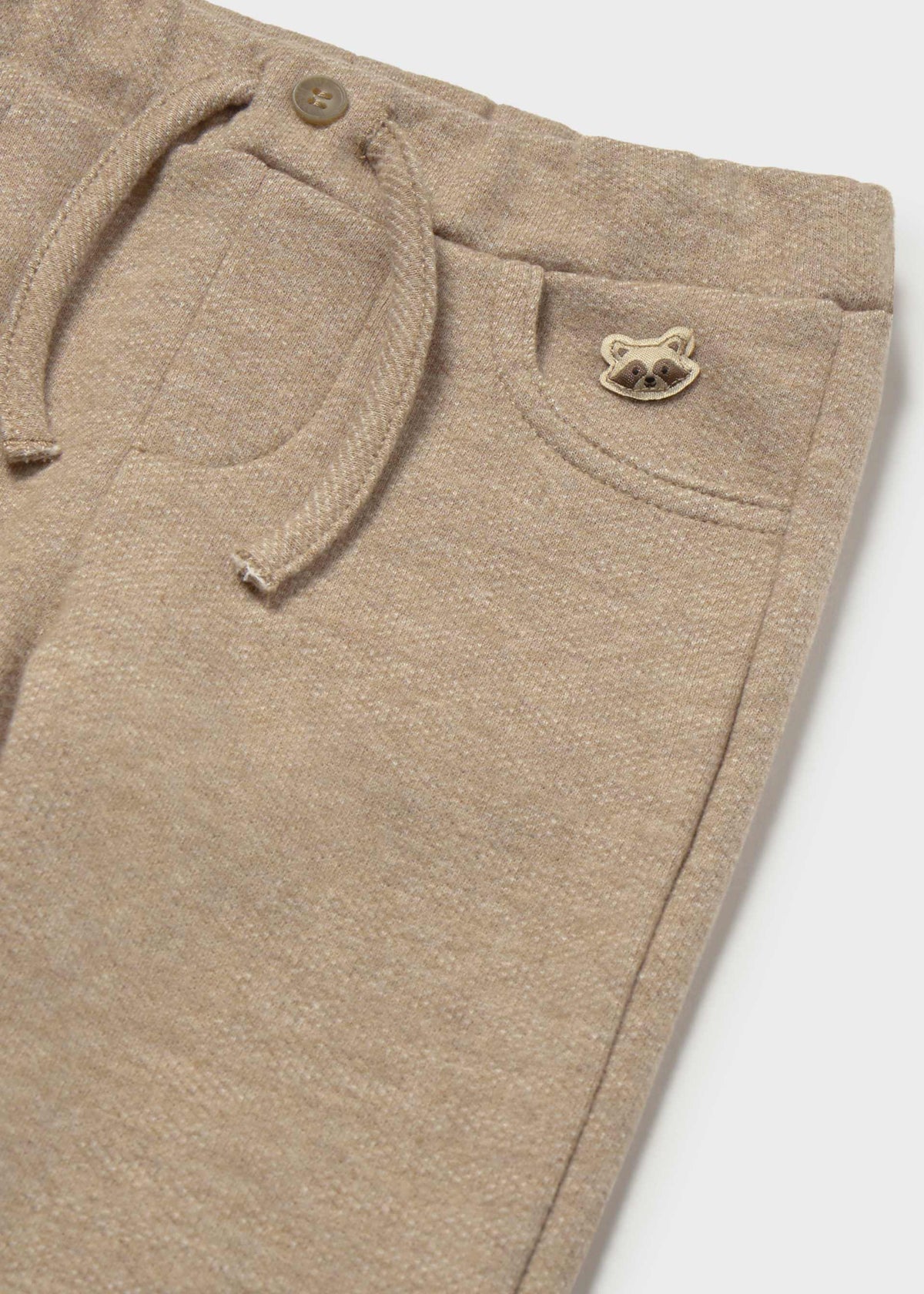 Close-up of beige heathered knit pants showing a drawstring waist, front pocket, and a small metallic raccoon logo on the pocket.