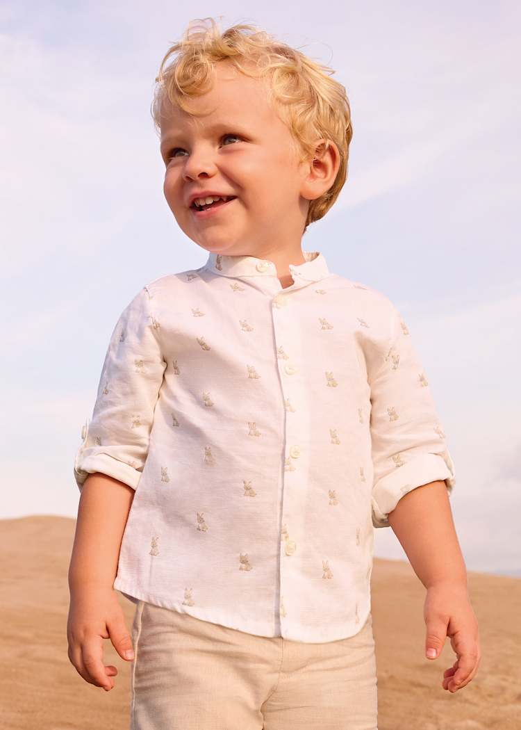 Young boy in a white shirt with a subtle pattern standing on sand with a clear sky.