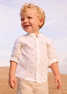 Young boy in a white shirt with a subtle pattern standing on sand with a clear sky.