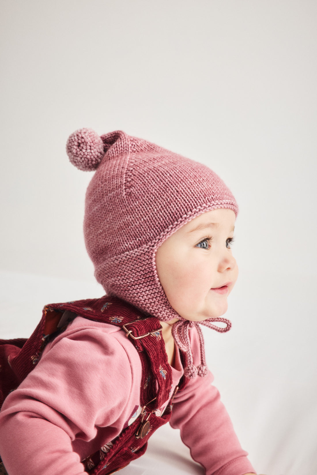 A baby wearing a dusty pink knit bonnet with ear flaps and a pom-pom, paired with a pink long-sleeve top and a dark red patterned overalls jacket, photographed against a light gray background.