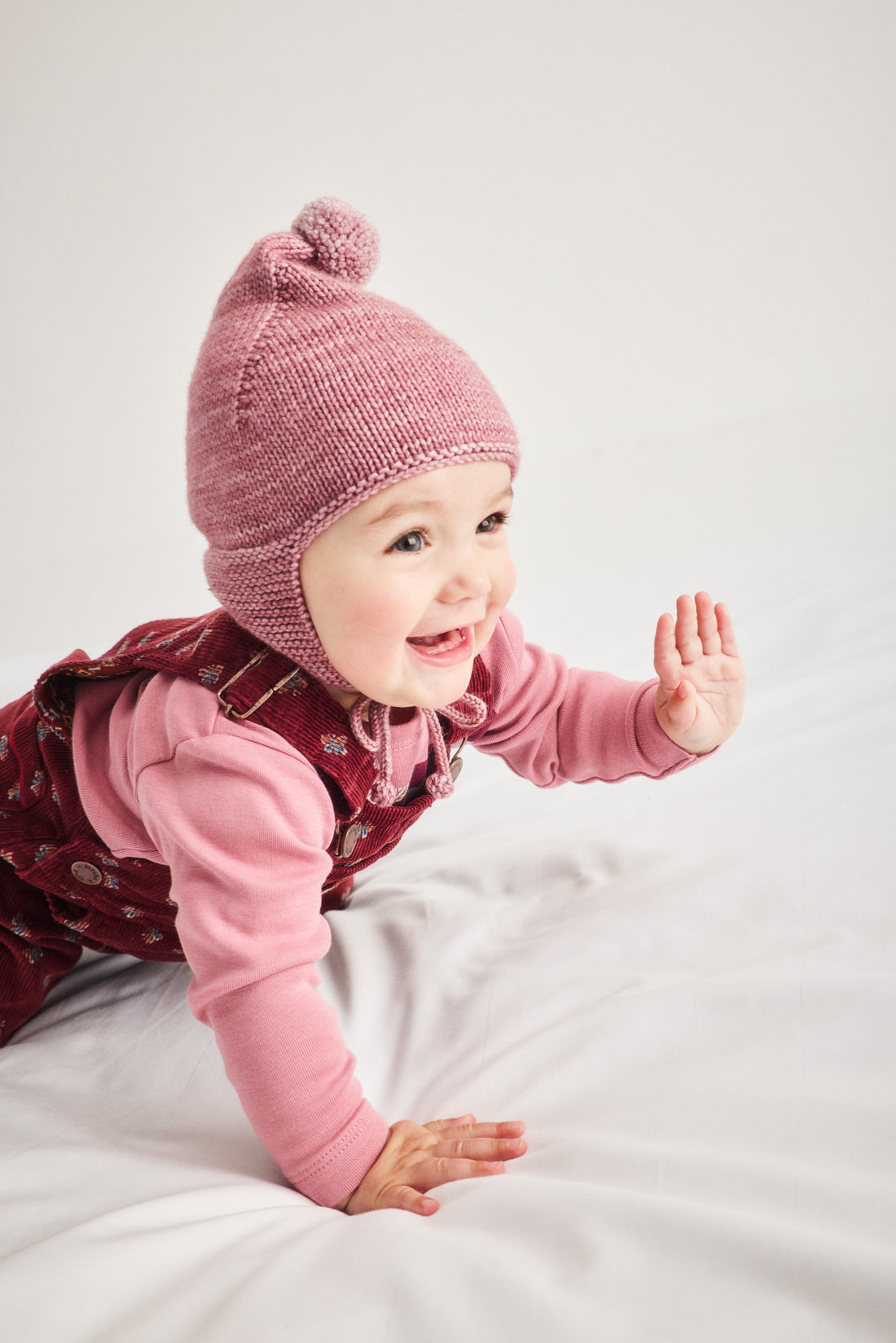 A smiling baby crawling on a white fabric surface, wearing a dusty pink knit pom-pom beanie and a pink long-sleeve top with a dark red patterned pinafore dress.