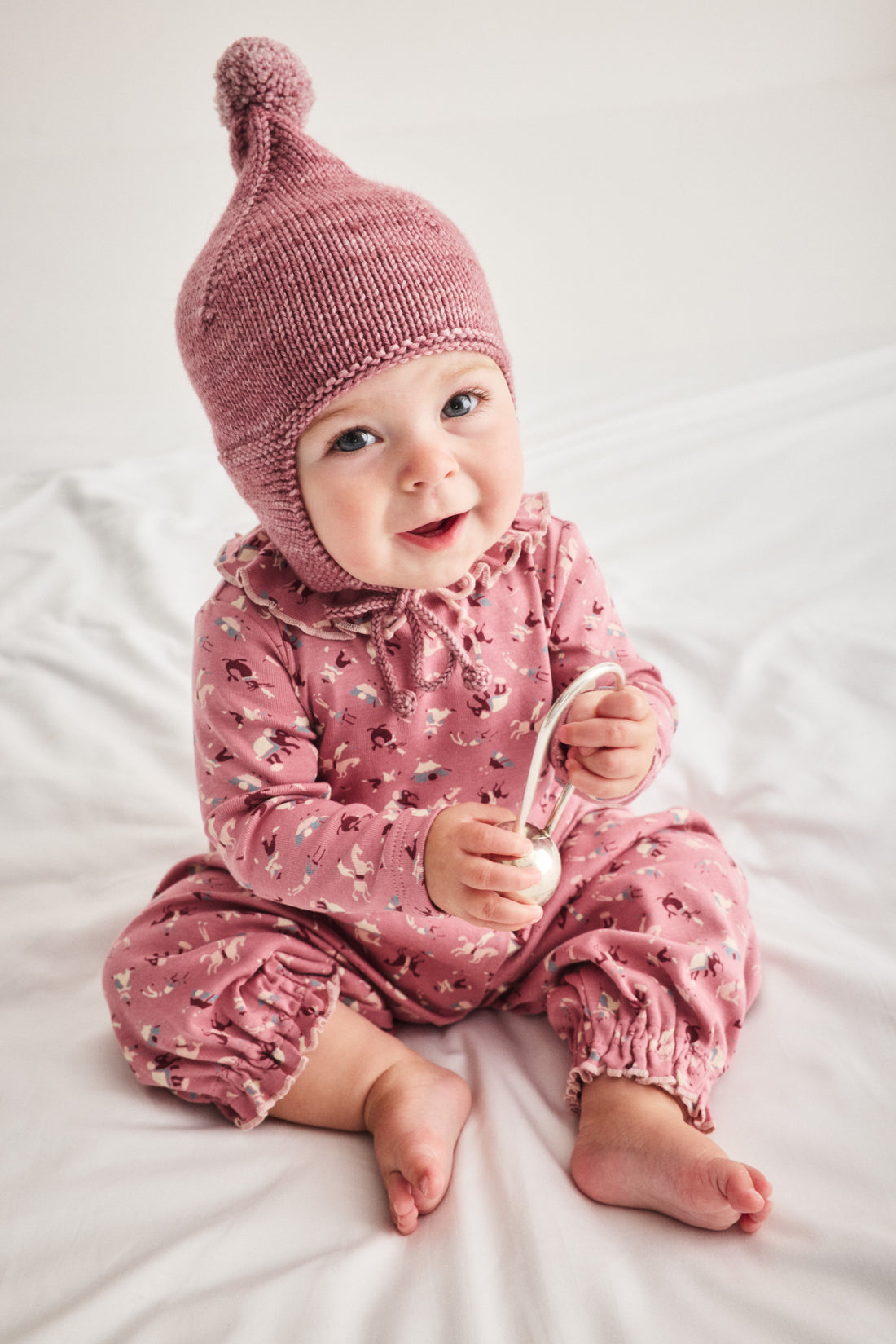 A baby sitting on white bedding wearing a dusty pink knit bonnet and matching pink patterned romper, holding a small white toy.
