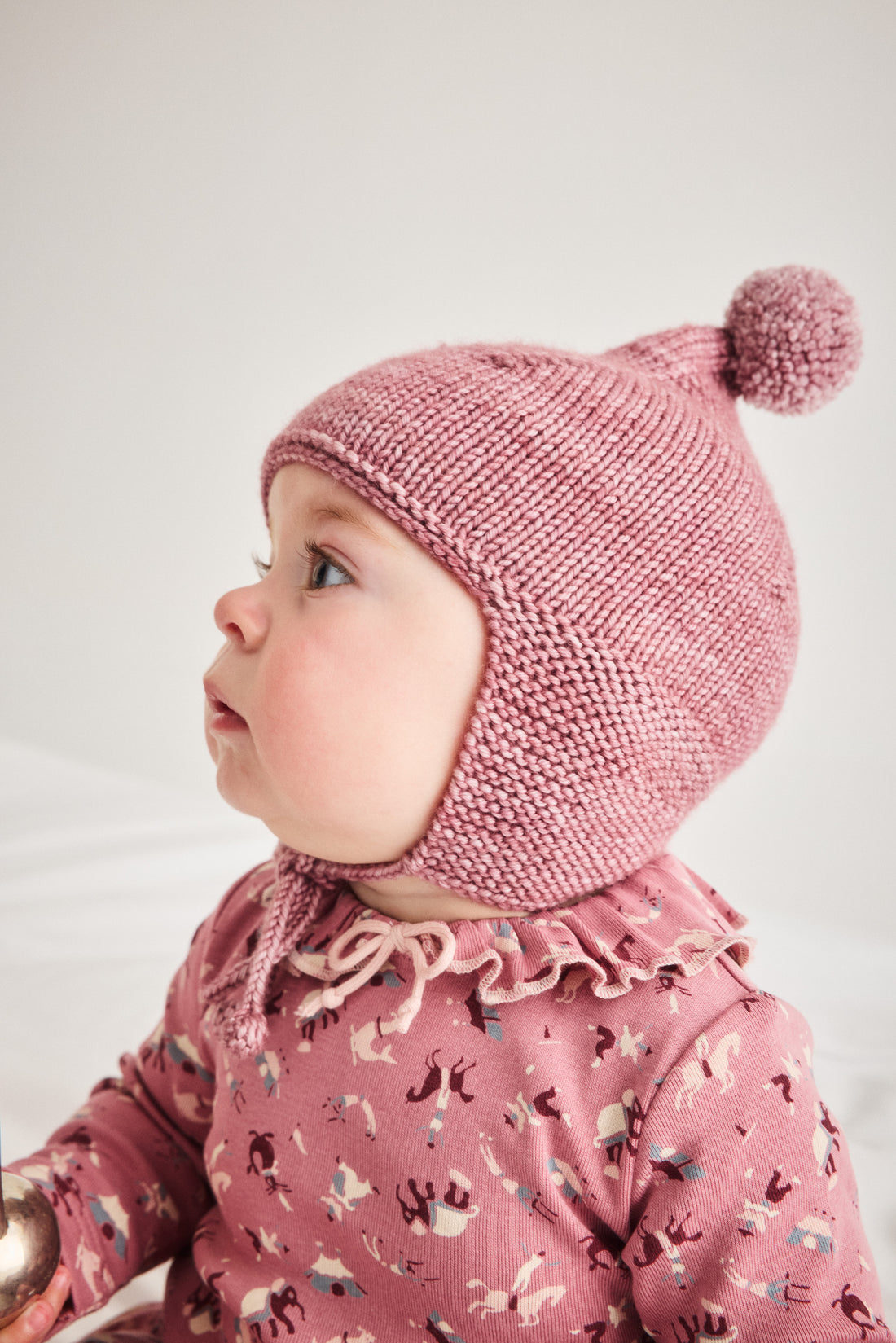 A baby wearing a dusty pink knit pom-pom bonnet and a matching pink patterned long-sleeve romper, shown in profile against a light neutral background.