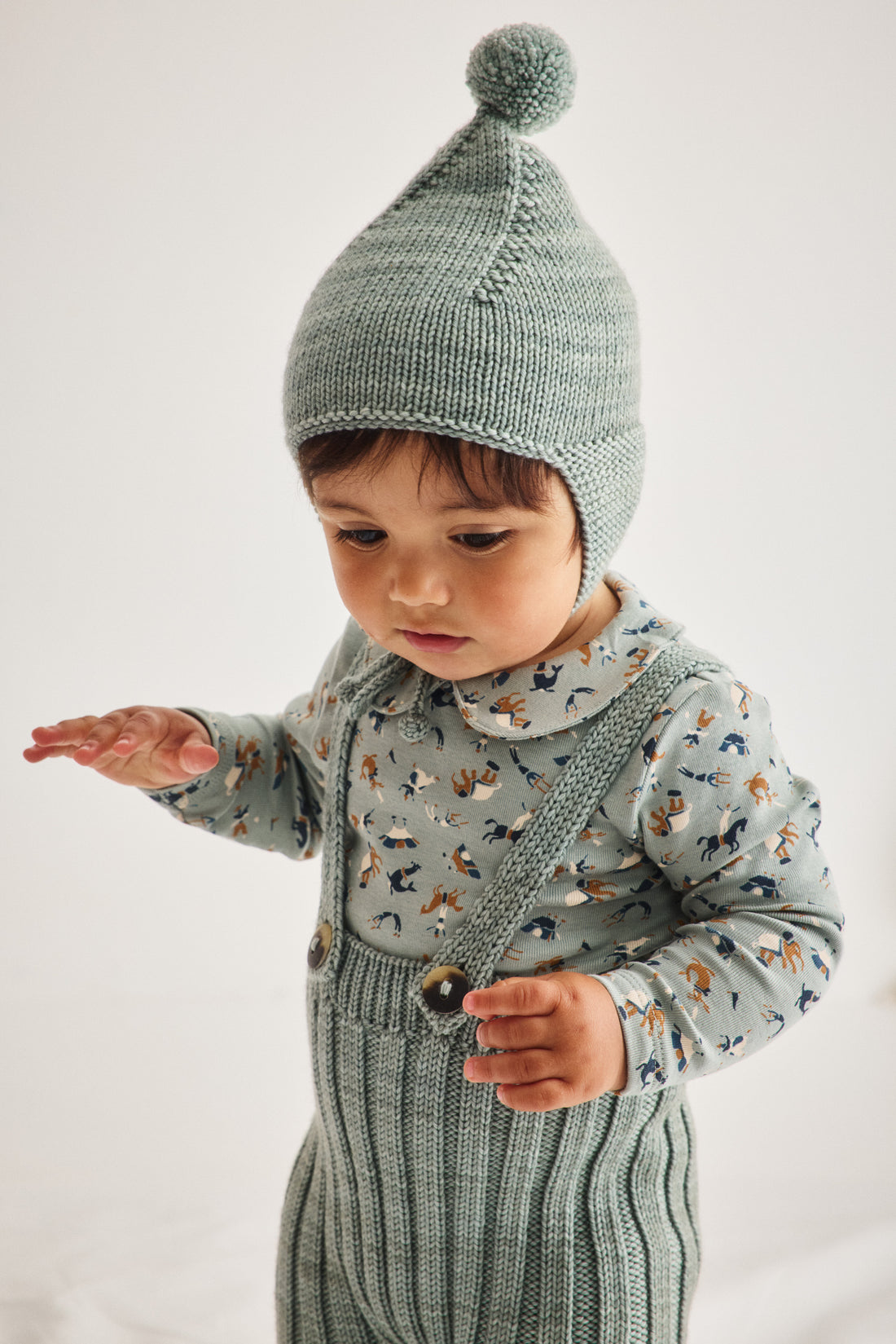 A toddler wearing a sage green knit bonnet with a pom-pom and a matching sage green ribbed overalls with a multicolor animal print, photographed against a plain light background.