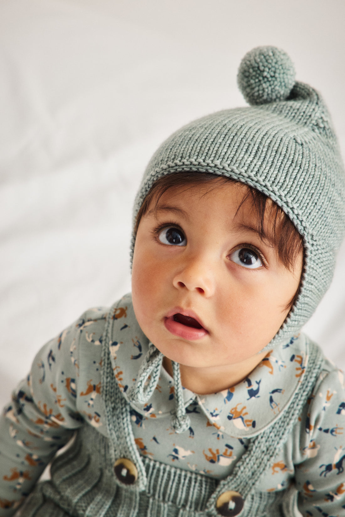 A close-up of a baby wearing a sage green knit bonnet with a pom-pom and a matching patterned long-sleeve top against a soft white background.