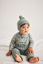 A baby sitting on white bedding wearing a sage green knit bonnet with a pom-pom, a matching patterned long-sleeve top, and green knit overalls.
