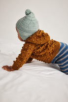 A baby crawling on white bedding, wearing a brown hooded knit sweater with a textured pom-pom pattern, a light blue knit beanie with a pom-pom, and blue striped pants.