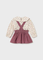 A toddler girl's two-piece outfit featuring a long-sleeve blouse with a cream floral print and a dusty rose smocked-elastic mini skirt with matching crossbody suspenders, laid flat on a light gray background.