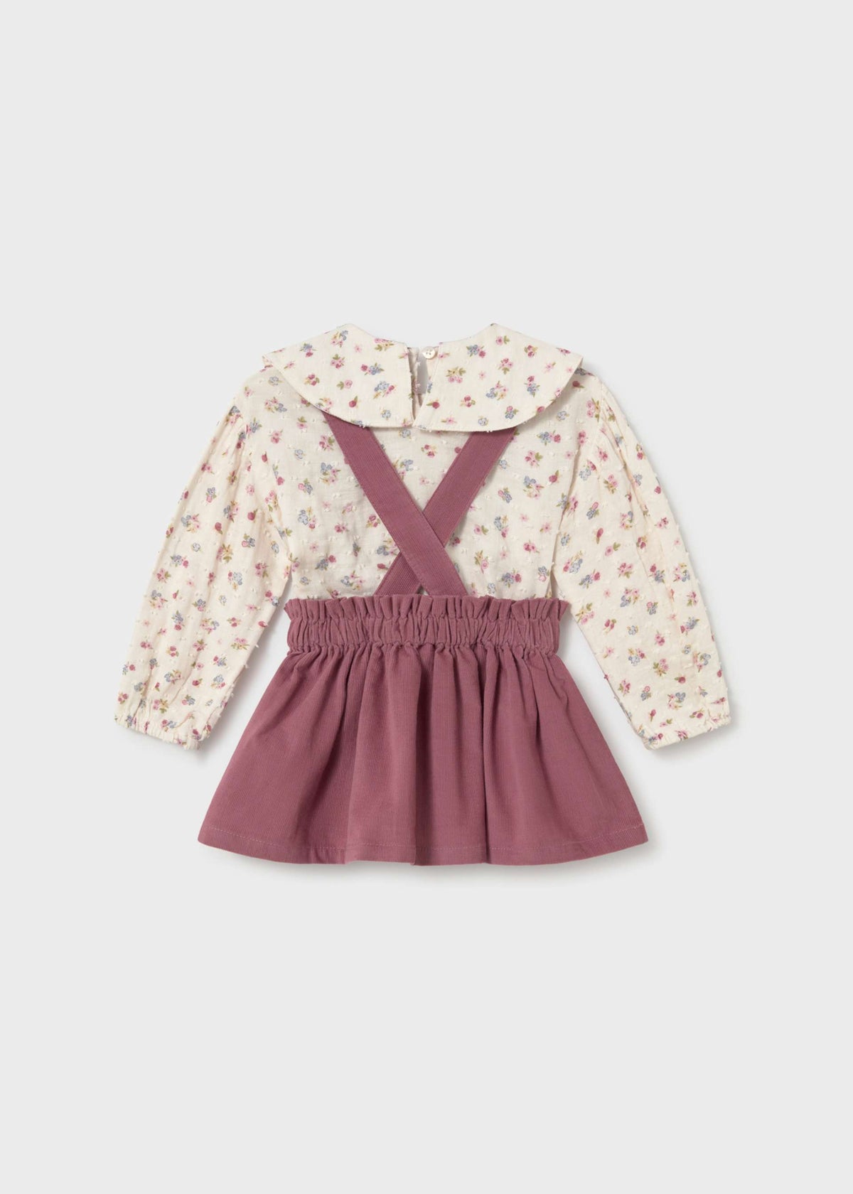 A toddler girl's two-piece outfit featuring a long-sleeve blouse with a cream floral print and a dusty rose smocked-elastic mini skirt with matching crossbody suspenders, laid flat on a light gray background.