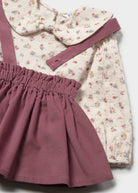 A children's outfit laid flat on a light gray background, featuring a cream floral long-sleeve top with a large bow collar and a dusty rose corduroy skirt with an elastic waistband.