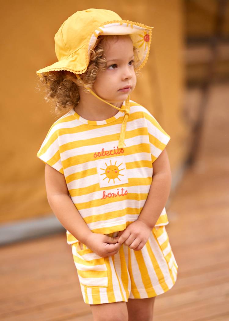 Child wearing a yellow and white striped outfit with a sun hat on a wooden floor.