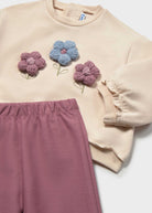 A children's two-piece outfit featuring a cream sweatshirt with three colorful crocheted flowers (pink, blue, and mauve) on the chest, paired with matching mauve sweatpants.