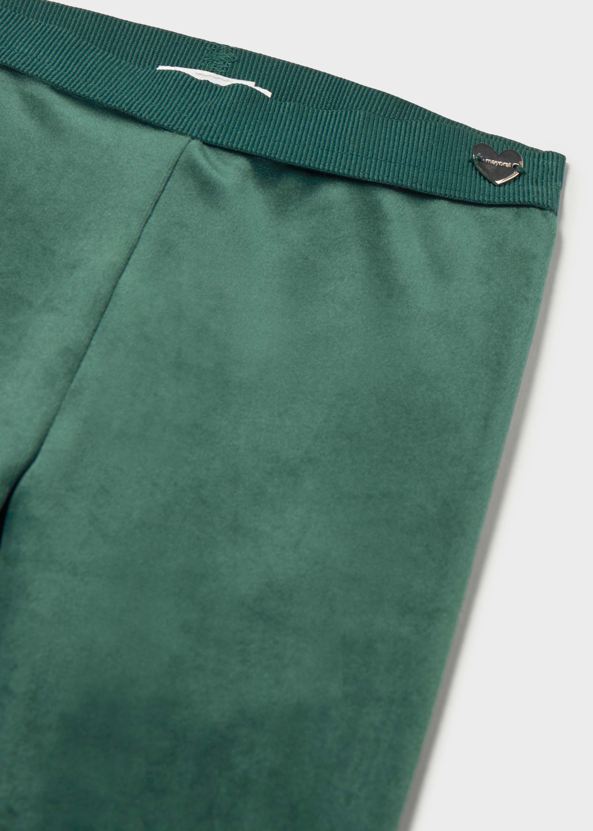 Close-up of green trousers showing the waistband with a white embroidered logo and a small metallic heart-shaped tag, on a light gray background.