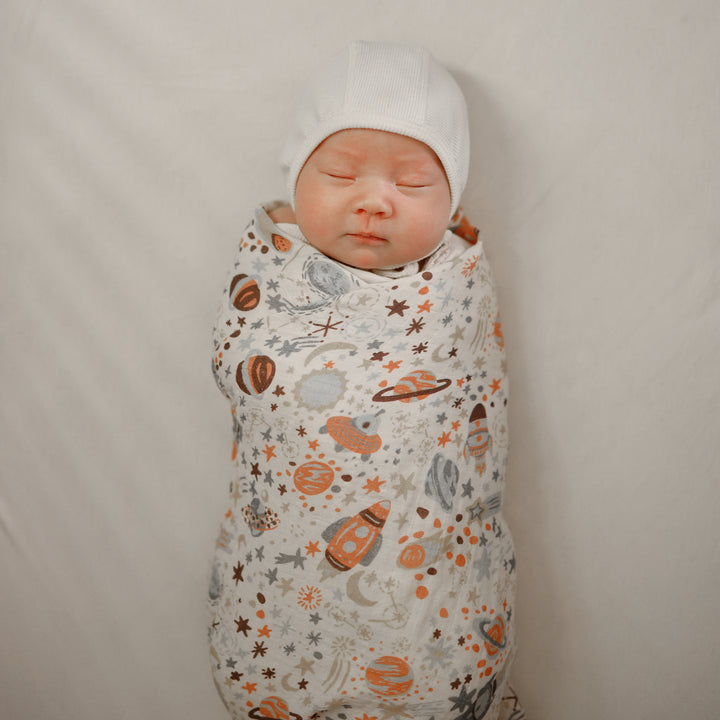 Newborn baby swaddled in a space-themed blanket with a white hat on a plain background