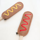 Two brown felt hot dog-shaped accessories on white sticks, one with yellow wavy embroidery and one with red wavy embroidery, on a white background.