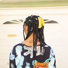 Person with braided hair wearing a patterned shirt in front of a white wall.