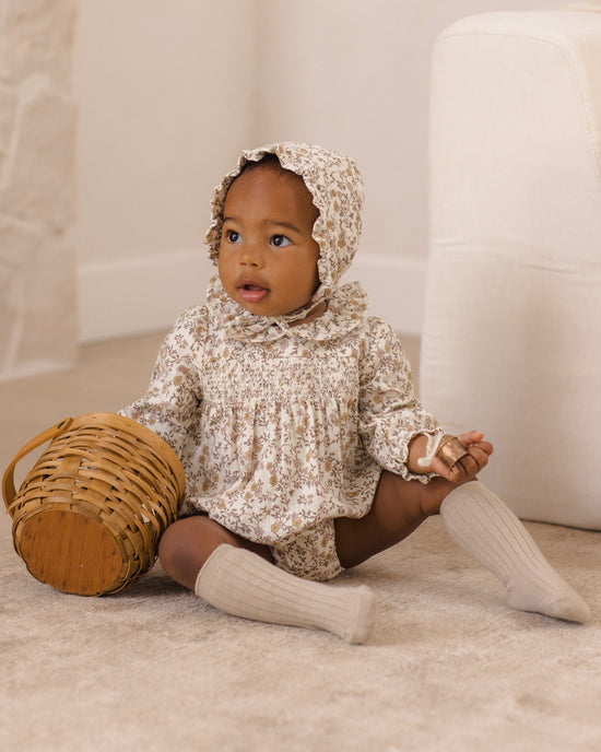 A baby sitting on a beige carpet wearing a cream and brown floral print smocked bodysuit with a matching bonnet, ribbed cream knee socks, and holding a small woven basket.