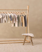 A wooden clothing rack holding a coordinated set of baby garments in soft neutral tones, including striped and solid tops, long-sleeve shirts, and pants, arranged in a styled studio setting with a small wooden stool and textured rug.