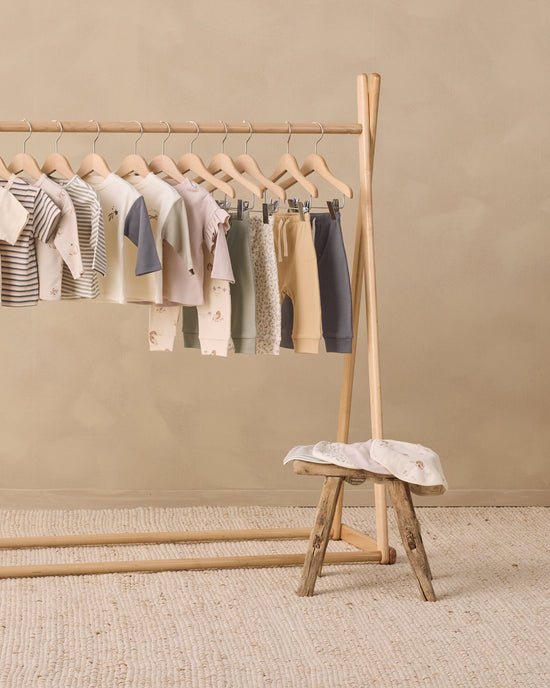 A wooden clothing rack holding a coordinated set of baby garments in soft neutral tones, including striped and solid tops, long-sleeve shirts, and pants, arranged in a styled studio setting with a small wooden stool and textured rug.