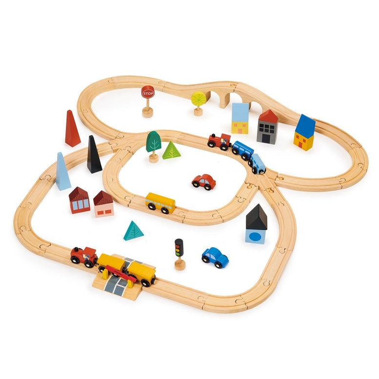 A close-up, angled view of a wooden toy train set featuring a red engine with a driver, a blue passenger car, and a yellow cargo car resting on wooden tracks. A round red "STOP" sign stands beside the track, and the background is filled with colorful wooden town elements, including a red house with a window, a yellow building, a blue bridge, and abstract green trees.