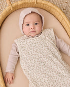 A baby wearing a cream floral-patterned sleeveless sleep sack over a light pink long-sleeve top and matching bonnet, lying on a beige cushion inside a woven rattan bassinet.