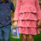 A child in a pink tiered long-sleeve dress holding a colorful 'Sea Life' book outdoors, standing next to another child in a navy blue shirt with blue dragonfly patterns.