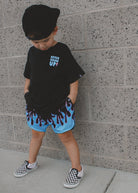 Child wearing a black t-shirt with text, blue shorts with red accents, and checkered shoes against a gray wall.