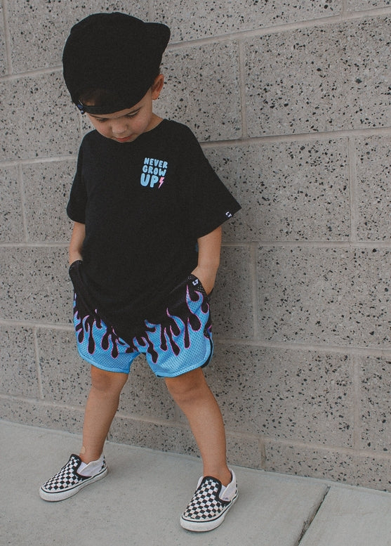 Child wearing a black t-shirt with text, blue shorts with red accents, and checkered shoes against a gray wall.