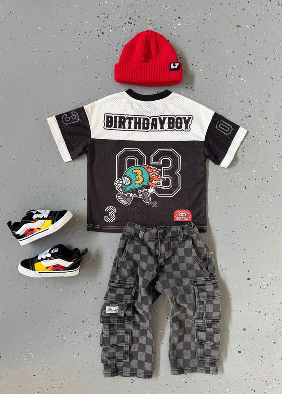 Children's birthday-themed outfit with shirt, pants, shoes, and red beanie on a gray surface.