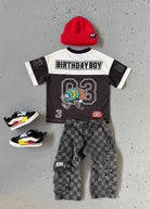 Children's birthday-themed outfit with shirt, pants, shoes, and red beanie on a gray surface.