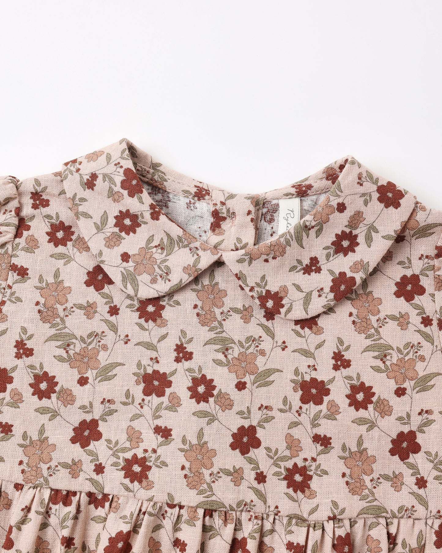 Close-up of a children's dress with a ruffled Peter Pan collar and a dusty floral print on a light beige linen-like fabric, featuring red and brown flowers with green leaves.