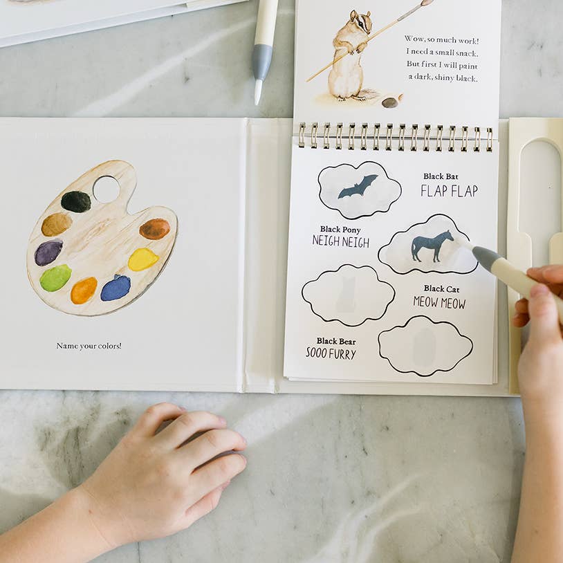 A child's coloring book shown open on a marble surface, with a hand holding a white paint pen. The left page shows a watercolor-style wooden paint palette, and the right page has animal-face cloud shapes for coloring, including Black Bear, Black Pony, Black Cat, and Black Car.