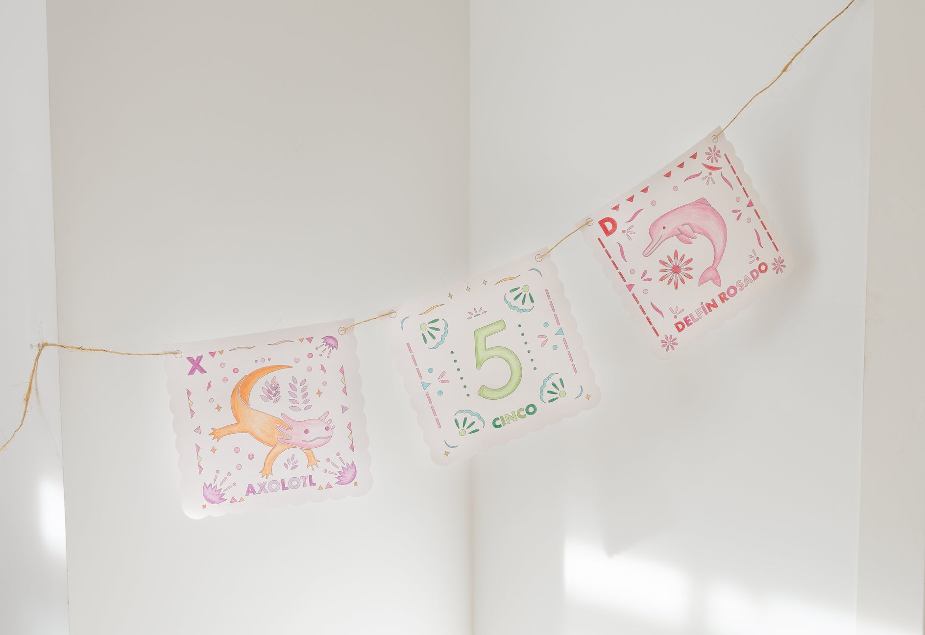 Three decorative square pennants strung along a thin cord, hanging against a white wall. The center pennant reads “5 Cinco” with green and orange lettering, while the side pennants feature pink and orange illustrations with Spanish text.